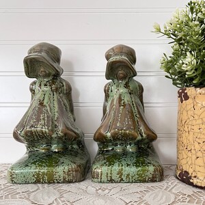 May include: A pair of green glazed ceramic bookends shaped like girls in dresses. The bookends are sitting on a white lace tablecloth with a yellow ceramic pot with a green plant in the background.
