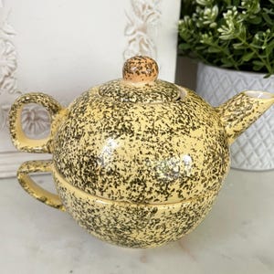 May include: A speckled yellow ceramic teapot with a matching cup. The teapot has a rounded shape, a spout, and a handle. The lid has a small, round knob. The cup fits neatly beneath the teapot. The speckled pattern is dark green.