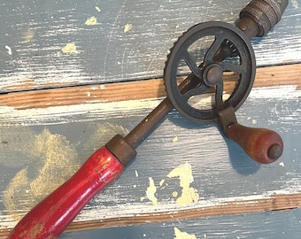 Puede incluir: Taladro manual antiguo con mango de madera rojo y marco de metal negro. El taladro cuenta con un sistema de engranajes y un portabrocas para sujetar brocas. El mango muestra signos de desgaste.