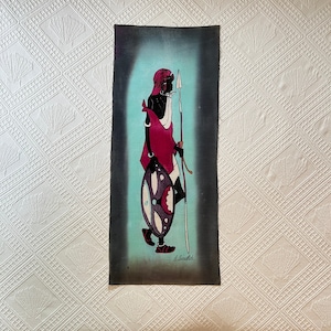 May include: A hand-painted batik wall hanging featuring a stylized figure holding a spear and shield. The figure is depicted in black with a pink and white patterned garment. The background is a gradient of blue and green.