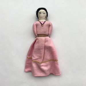 May include: A handmade fabric doll wearing a pink dress with gold trim and a brown belt. The doll has black hair, a painted face, and beaded earrings and necklace.
