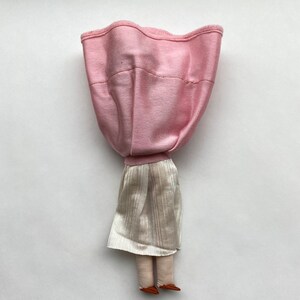 May include: A handmade doll with a pink fabric dress and a white skirt. The doll has brown fabric legs and feet.