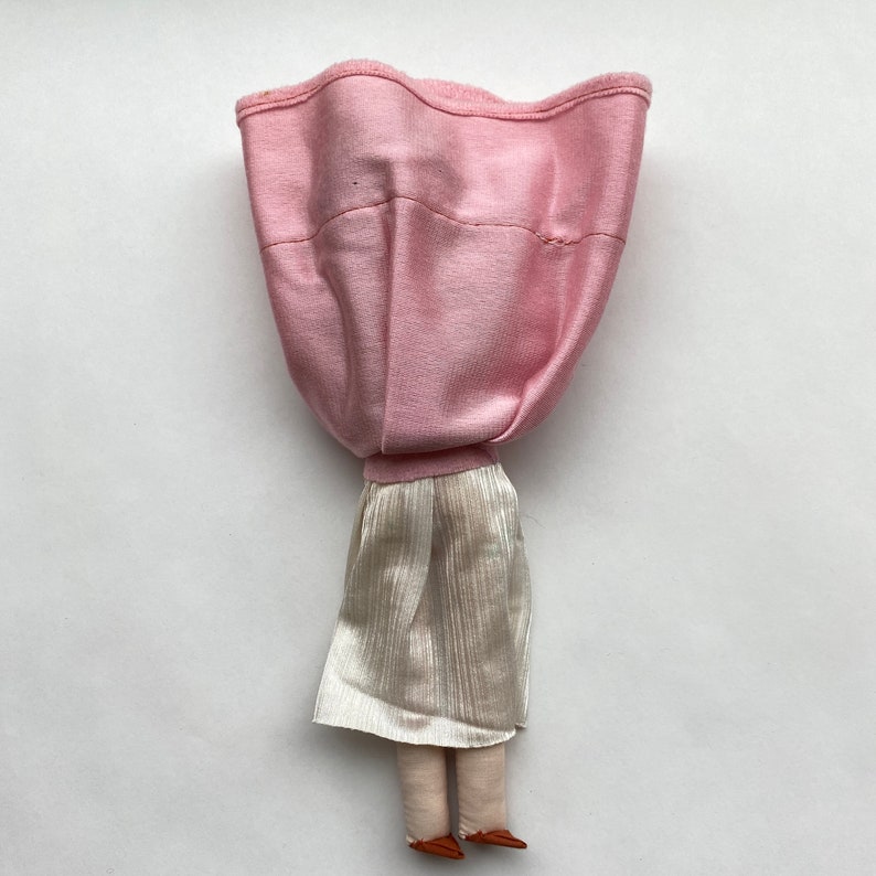 May include: A handmade doll with a pink fabric dress and a white skirt. The doll has brown fabric legs and feet.