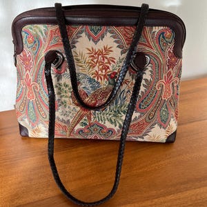 以下が含まれることがあります： ペイズリーとキジのプリントが施された、茶色の革と布製のトートバッグ。バッグには茶色の革のストラップが2本付いています。