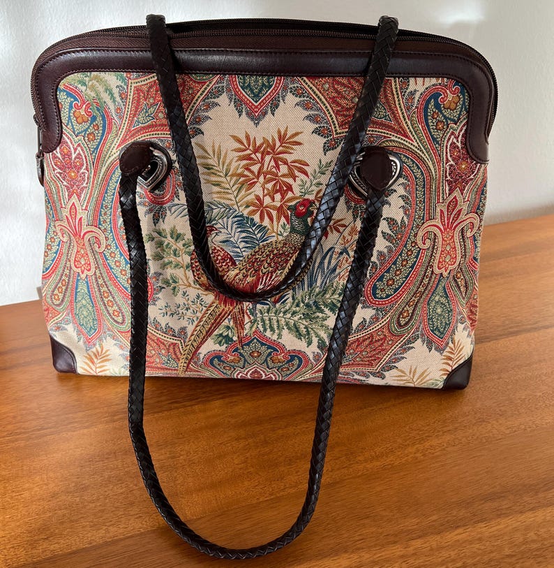 以下が含まれることがあります： ペイズリーとキジのプリントが施された、茶色の革と布製のトートバッグ。バッグには茶色の革のストラップが2本付いています。