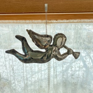 May include: A silver angel ornament with wings outstretched, holding a trumpet in its hand. The ornament is hanging from a chain against a white lace curtain background.