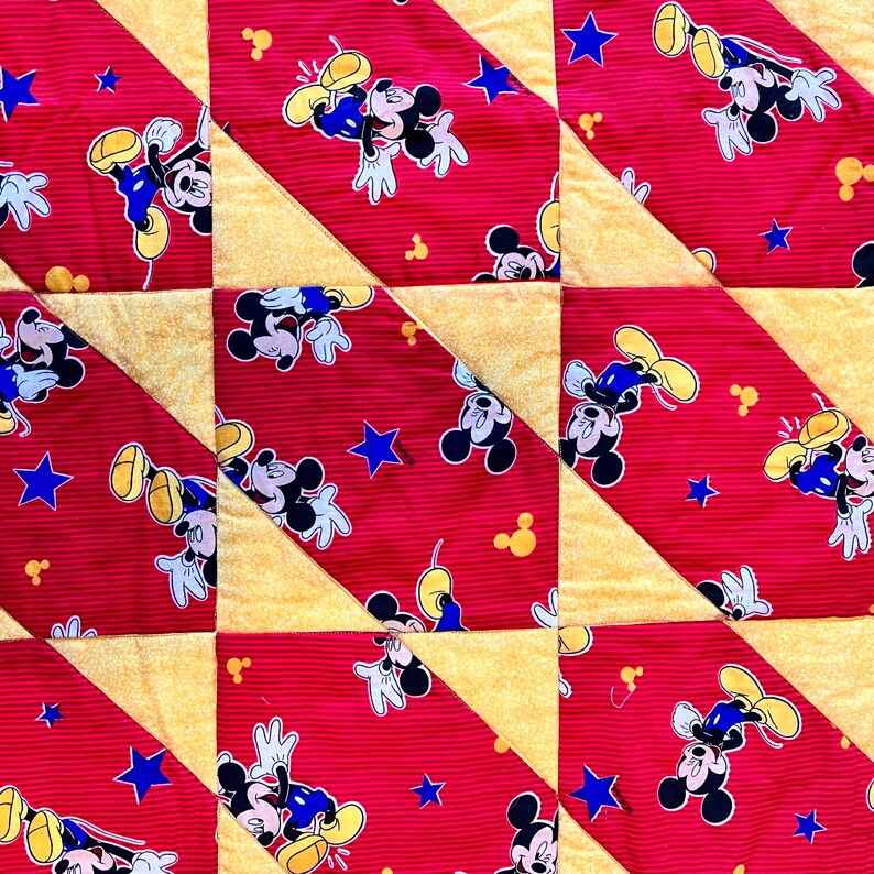 May include: A patchwork quilt featuring a repeating pattern of Mickey Mouse on a red striped background. The design includes blue stars and yellow triangles. The quilt is likely handmade and suitable for a child's room.