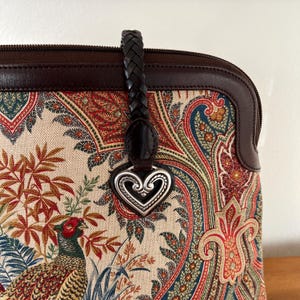 以下が含まれることがあります： ブラウンレザーとペイズリー柄のハンドバッグに、シルバーのハート型のチャームが付いています。バッグには編み込みのレザーストラップが付いています。