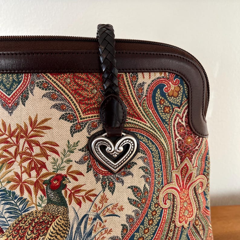 以下が含まれることがあります： ブラウンレザーとペイズリー柄のハンドバッグに、シルバーのハート型のチャームが付いています。バッグには編み込みのレザーストラップが付いています。
