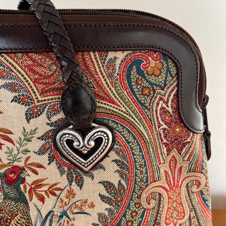 以下が含まれることがあります： ブラウンレザーとペイズリー柄のハンドバッグに、シルバーのハート型のチャームが付いています。バッグには編み込みのレザーストラップが付いています。