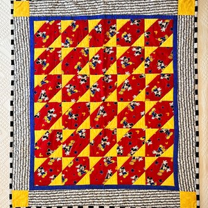 May include: A handmade quilt featuring a repeating pattern of Mickey Mouse on a red background with yellow and blue accents. The quilt has a black and white striped border with yellow corner squares. The quilt is likely made of cotton.