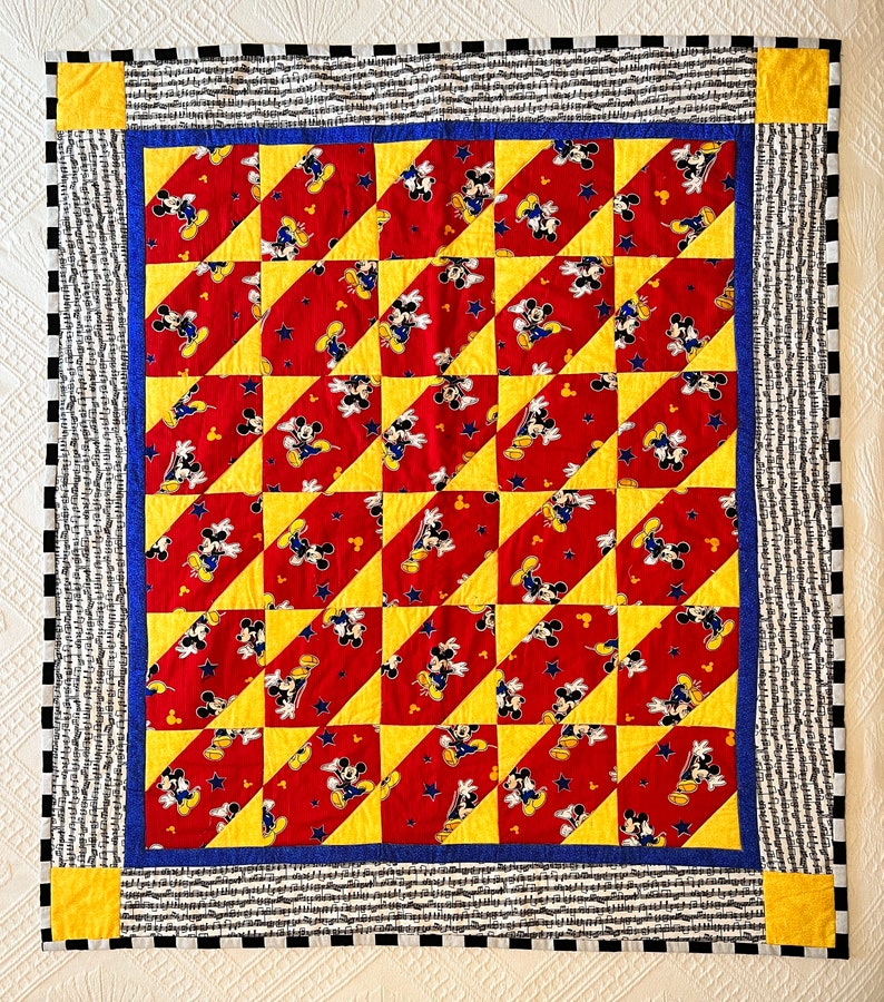 May include: A handmade quilt featuring a repeating pattern of Mickey Mouse on a red background with yellow and blue accents. The quilt has a black and white striped border with yellow corner squares. The quilt is likely made of cotton.