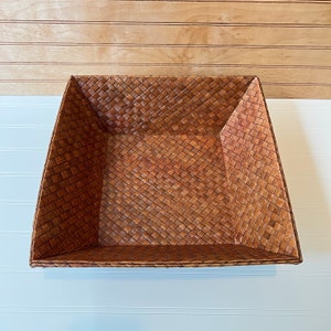 May include: A square woven basket made of natural brown material. The basket has a woven pattern and is empty.
