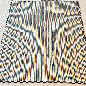 May include: A colorful quilt with vertical stripes in yellow, green, purple, and navy blue. The quilt has a white background and a black and white checkered border. The stripes are evenly spaced and run the length of the quilt.