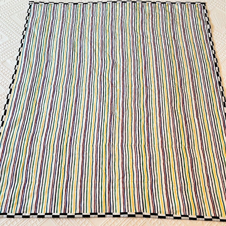 May include: A colorful quilt with vertical stripes in yellow, green, purple, and navy blue. The quilt has a white background and a black and white checkered border. The stripes are evenly spaced and run the length of the quilt.
