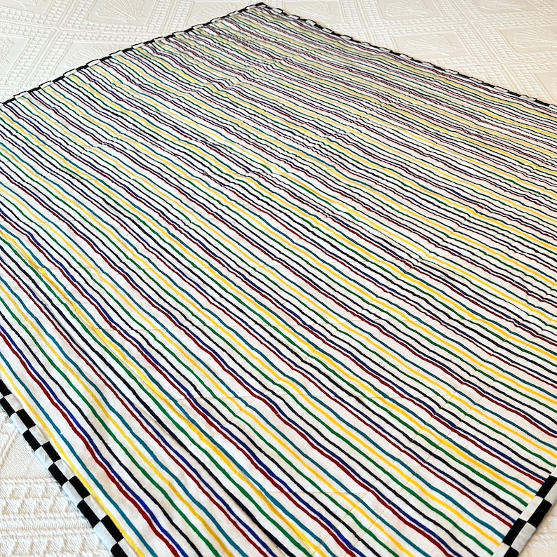 May include: A colorful quilt with a white background and vertical stripes in red, yellow, green, blue, and black. The quilt has a black and white checkered border on one side.