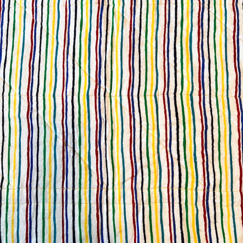 May include: A close-up view of a quilted fabric with vertical stripes in various colors. The stripes include red, yellow, green, blue, and black, set against a white background. The quilting pattern features diagonal stitching.