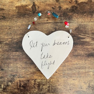 May include: White ceramic heart-shaped ornament with the text "Let your dreams take flight" written in black ink. The ornament is hanging from a string with colorful beads.