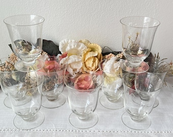 May include: Set of eight clear glass wine glasses with a delicate design. The glasses are arranged in a circle on a white tablecloth with pink and yellow flowers.
