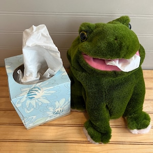 May include: A plush green frog tissue holder with a pink mouth and white tissue sticking out. Next to it is a blue tissue box with a floral design and white tissues coming out of the top.