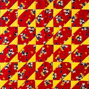 May include: A patchwork quilt featuring a repeating pattern of Mickey Mouse on a red background with yellow and blue accents. The quilt is made of fabric with a ribbed texture, and the design includes stars and Mickey Mouse's gloves.