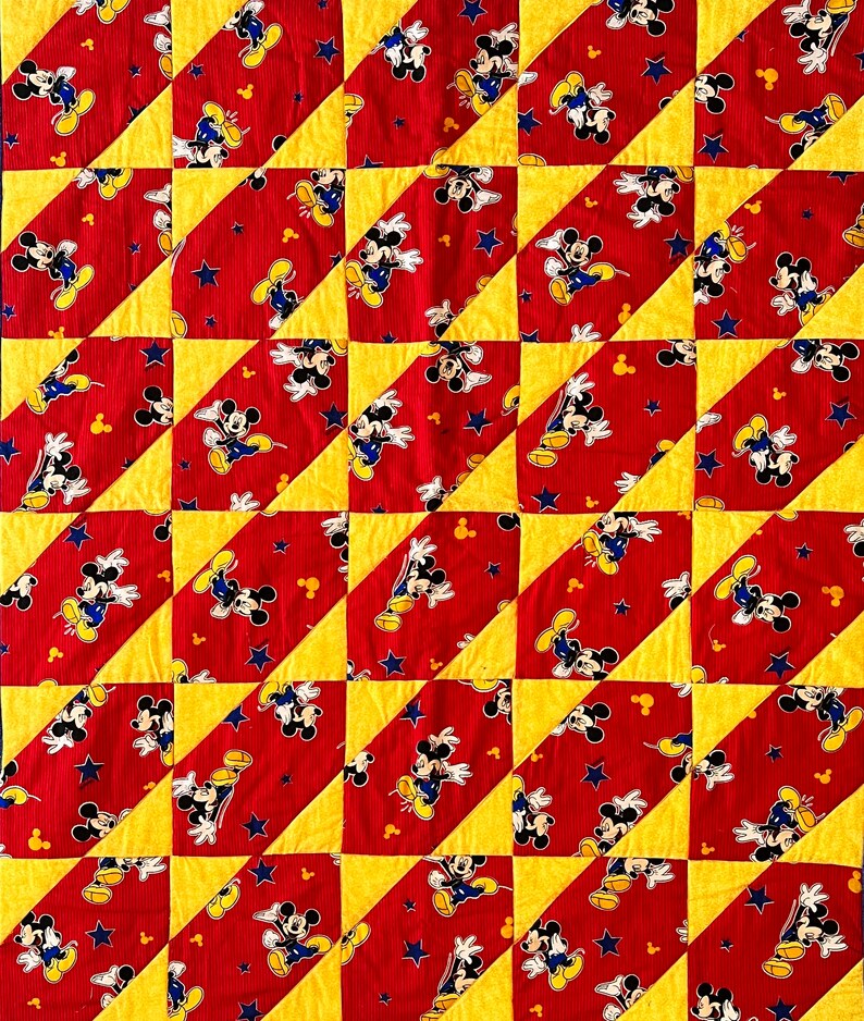 May include: A patchwork quilt featuring a repeating pattern of Mickey Mouse on a red background with yellow and blue accents. The quilt is made of fabric with a ribbed texture, and the design includes stars and Mickey Mouse's gloves.