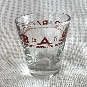 May include: Clear shot glass with a red design. The design includes the letters "B" and "A" with horseshoe and rope accents. The shot glass is made of clear glass and has a slightly angled rim.