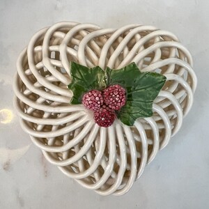 May include: Cream-colored ceramic heart-shaped basket with a woven design. The basket is adorned with three red berries and two green leaves. This decorative piece is suitable for displaying small items or as a unique accent.