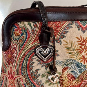 以下が含まれることがあります： ペイズリーと花のプリントが施された茶色の革製のハンドバッグ。バッグには編み込みの革のストラップと銀色のハートのチャームが付いています。