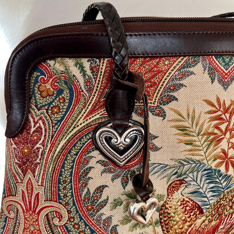 以下が含まれることがあります： ペイズリーと花のプリントが施された茶色の革製のハンドバッグ。バッグには編み込みの革のストラップと銀色のハートのチャームが付いています。