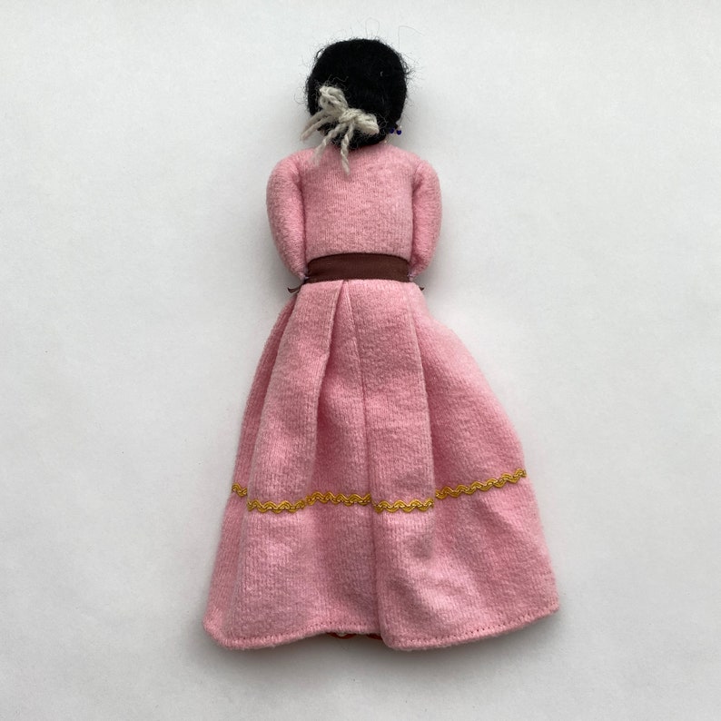 May include: A pink fabric doll with black hair, a brown belt, and gold trim on the skirt.