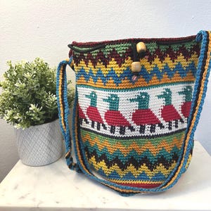 May include: A colorful, handmade crochet shoulder bag. The bag features a vibrant pattern with ducks in red dresses and teal heads. The bag has a blue, yellow, and black strap and wooden toggle closure. The bag is sitting on a white surface.
