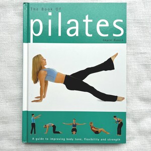 May include: A book titled "The Book of Pilates" with a teal and white cover. The cover features a woman in athletic wear demonstrating a Pilates pose. Additional images of Pilates exercises are at the bottom. The text reads "A guide to improving body tone, flexibility and strength."