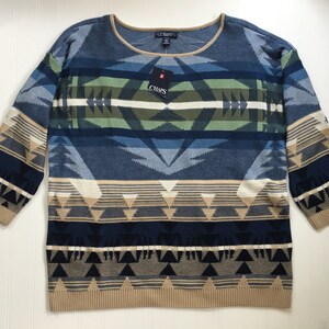 May include: A blue and brown patterned sweater with a crew neck. The sweater has a geometric design with a tan, blue, green, and white color scheme. The sweater has the word "Chaps" on the tag.