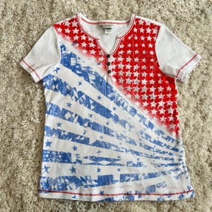 May include: White short-sleeve shirt with a V-neck and a patriotic design. The shirt features a red and white star pattern on the right side and blue and white stripes on the left side. The shirt has a button-up placket and red stitching.