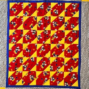 May include: A handmade quilt featuring a repeating pattern of Mickey Mouse on a red background with yellow and blue accents. The quilt has a blue border and a striped outer border. The quilt is a square shape.