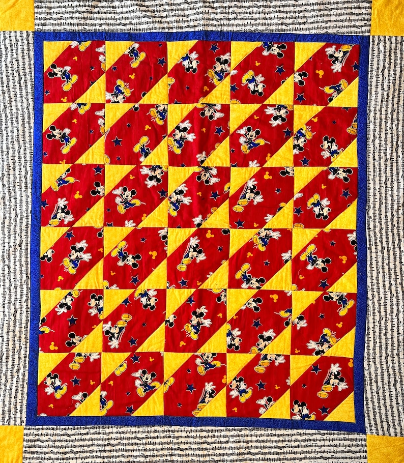 May include: A handmade quilt featuring a repeating pattern of Mickey Mouse on a red background with yellow and blue accents. The quilt has a blue border and a striped outer border. The quilt is a square shape.