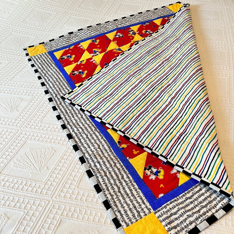 May include: A colorful quilt with a Mickey Mouse theme. The quilt features red, yellow, blue, and black accents. The front has a Mickey Mouse pattern, while the back has a striped design. The border is black and white.