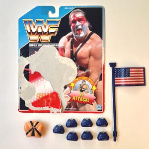 May include: A vintage WWF wrestling action figure set featuring a muscular wrestler with face paint. The set includes an American flag, blue accessories, and a torso piece. The packaging is partially torn.