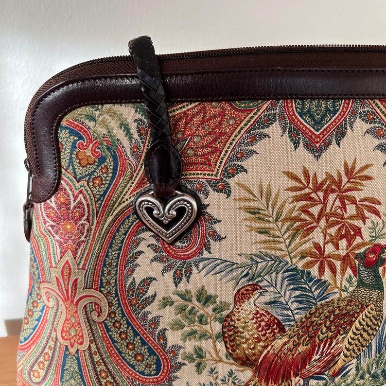 以下が含まれることがあります： ハート型のチャームが付いた、ペイズリー柄のブラウンレザーとキャンバスのハンドバッグ。バッグにはトップハンドルとショルダーストラップが付いています。