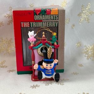 May include: A red, white, and blue Christmas ornament featuring a nutcracker doll in a clock tower. The ornament is in its original box, which reads "The Trimmerry Ornaments".