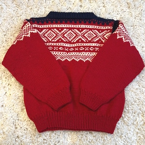 May include: A red knit sweater with white geometric patterns across the chest and shoulders. The sweater has a navy blue collar and cuffs. The sleeves are long and the material appears to be a thick knit. The sweater is laying flat on a white, textured surface.