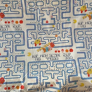 May include: White fabric with a blue Pac-Man maze pattern. The design includes Pac-Man characters, ghosts, and fruit. Text includes "PAC-MAN", "1UP HIGH SCORE", and score numbers. A fun, retro video game theme.