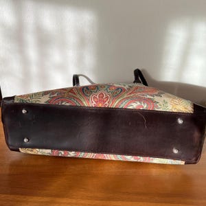 以下が含まれることがあります： ペイズリー柄のファブリックの外側に、ブラウンのレザーのトリミングが施されたトートバッグ。バッグはトップハンドルとジッパーの留め具が付いています。