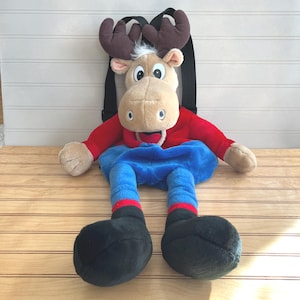 May include: A plush moose backpack with a tan face, brown antlers, and a red shirt. The backpack has blue pants with red stripes and black feet. The backpack straps are black.