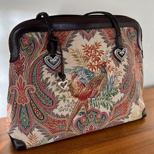 以下が含まれることがあります： 2つの銀色のハート形のチャームが付いた、ブラウンレザーとペイズリー柄のトートバッグ。バッグはキジのデザインです。