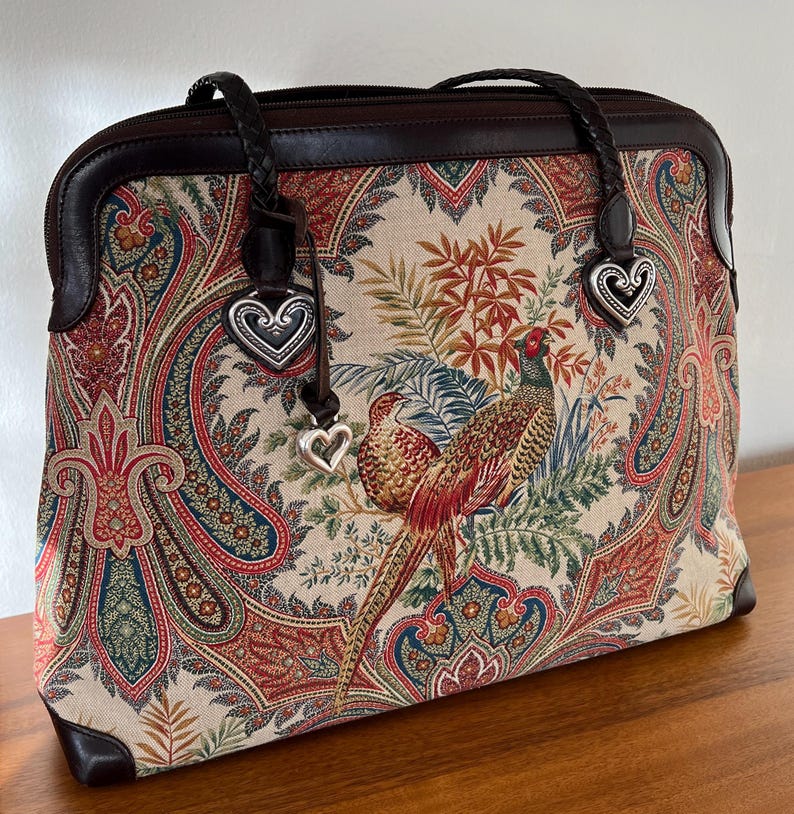 以下が含まれることがあります： 2つの銀色のハート形のチャームが付いた、ブラウンレザーとペイズリー柄のトートバッグ。バッグはキジのデザインです。