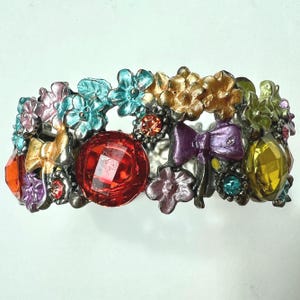 May include: A colorful bracelet with a floral design. The bracelet features various colored flowers, bows, and gemstones. The gemstones are red, orange, and yellow. The flowers are pink, blue, and gold.