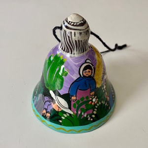 May include: A hand-painted ceramic bell with a purple, green, and white design. The bell features a scene of people in a garden with flowers and trees. The bell has a black cord for hanging.