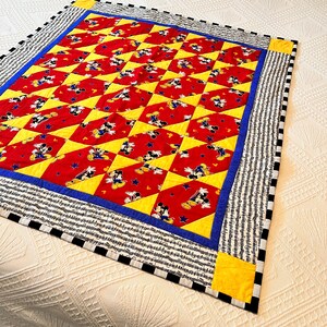May include: A handmade quilt featuring a repeating pattern of Mickey Mouse on a red background, interspersed with yellow diamond shapes. The quilt has a blue border and a black and white striped edge.
